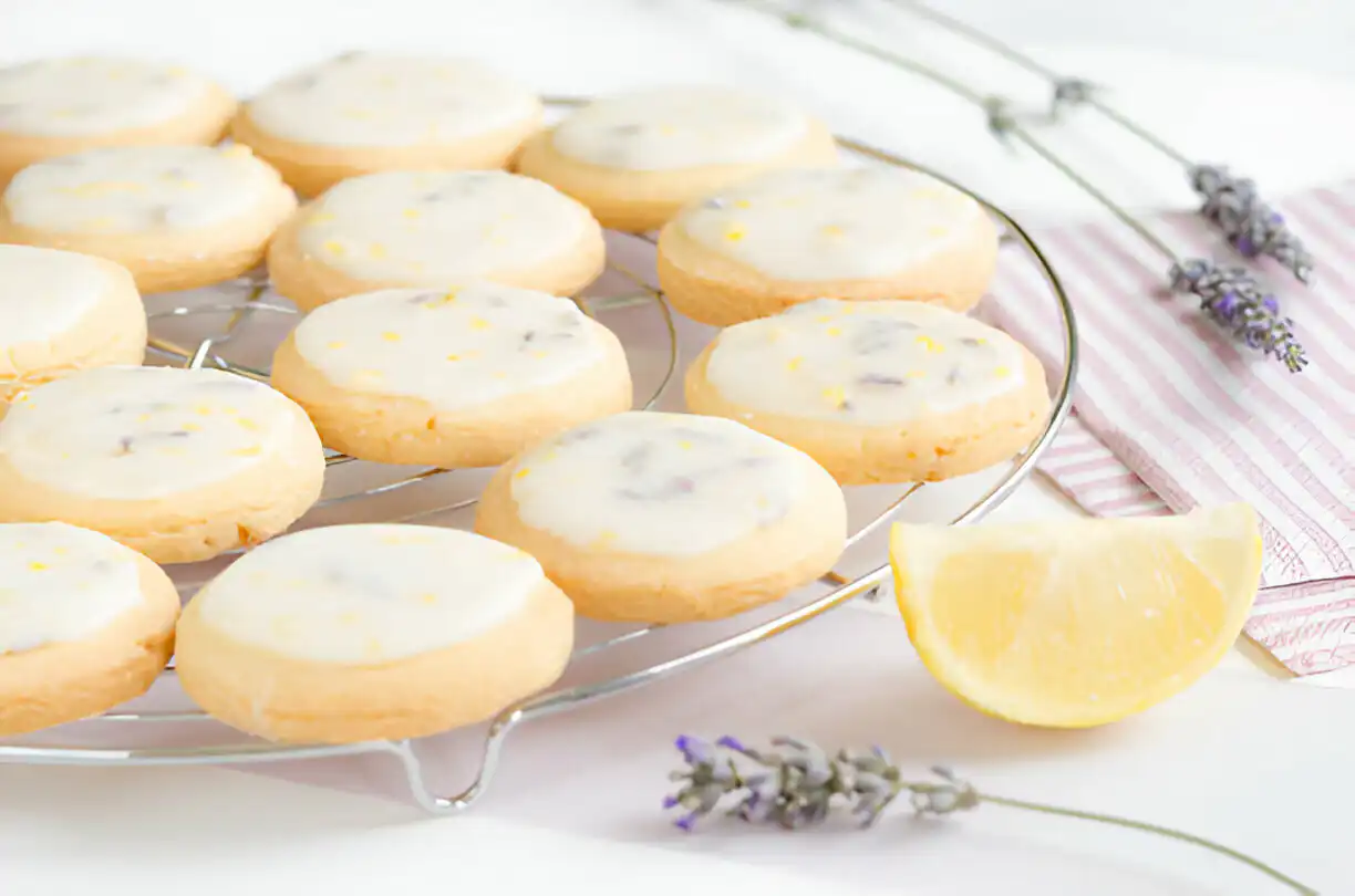 A plate of lemon lavender sugar cookies cooling on a wire rack with a slice of lemon and lavender sprigs nearby.