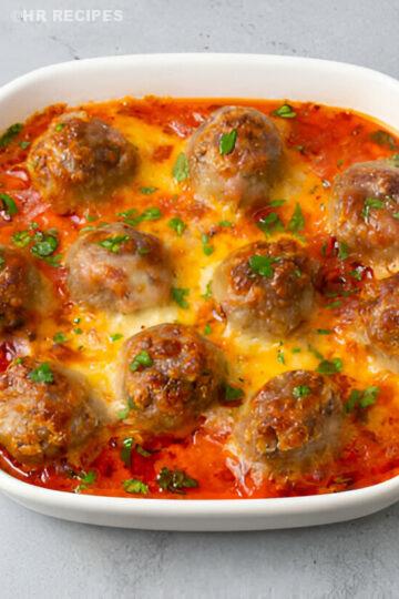 A dish of meatballs in tomato sauce topped with melted cheese and parsley, served in a white baking dish